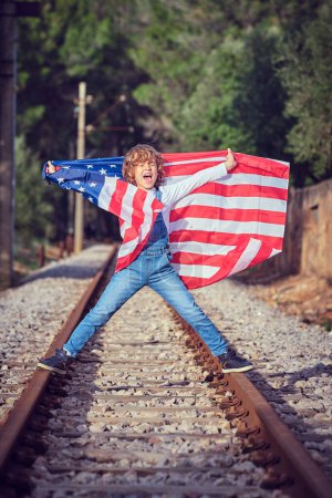 Téléchargez les photos : Corps complet de joyeux garçon préadolescent debout sur des rails de chemin de fer avec le drapeau des États-Unis d'Amérique et criant joyeusement - en image libre de droit