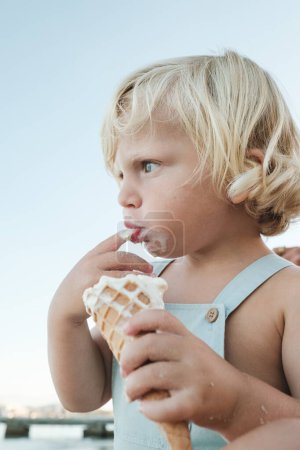 Foto de Lindo niño comiendo delicioso helado en cono de gofre y mirando hacia otro lado mientras está sentado en la playa - Imagen libre de derechos
