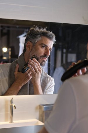 Foto de Hombre serio con toalla mirando a la reflexión en el espejo y barba de recorte en el baño moderno en casa - Imagen libre de derechos