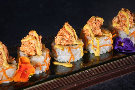 Téléchargez les photos : Gros plan de sushis appétissants garnis de saumon et de sauce fraîche servis sur un plateau en bois avec des fleurs au restaurant sur fond noir - en image libre de droit