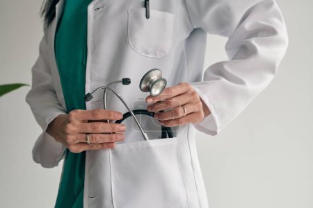 Téléchargez les photos : Cultiver médecin femme méconnaissable en uniforme médical mettre stéthoscope dans la poche sur fond clair - en image libre de droit