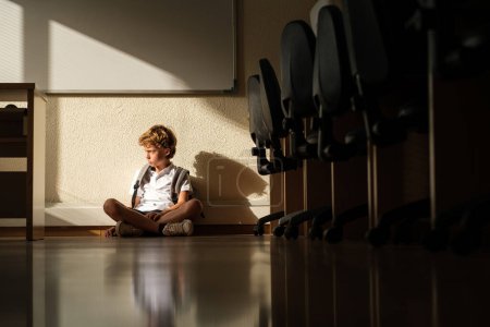 Téléchargez les photos : Offensé écolier fronçant les sourcils et assis seul sur le sol dans la salle de classe tout en regardant loin - en image libre de droit