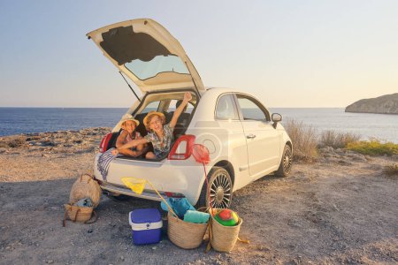 Téléchargez les photos : Deux enfants assis dans le coffre de la voiture avec des gestes de liberté avec des paniers d'objets pour la plage et une glacière portable à côté de la voiture avec la mer en arrière-plan - en image libre de droit