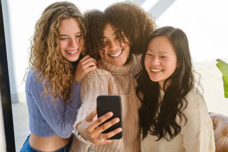 Photo for Happy young diverse women standing close to each other smiling and taking selfie on smartphone - Royalty Free Image