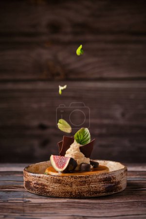 Foto de Proceso de decoración de pastel dulce rematado con hojas de menta crema batida chocolate y rebanada de higo y servido en plato de cerámica en mesa de madera - Imagen libre de derechos