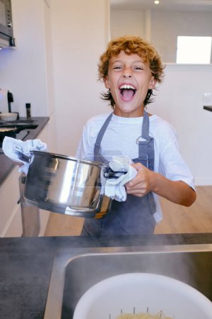 Téléchargez les photos : Enfant heureux dans un tablier avec pot regardant caméra contre évier avec des pâtes en passoire à la maison - en image libre de droit