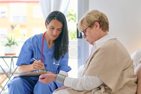 Photo for Female doctor in uniform with clipboard against senior patient with digital blood pressure monitor on wrist during checkup in house - Royalty Free Image