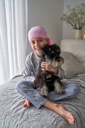 Foto de Cuerpo completo de niño sonriente descalzo en pijama y bandana rosa como símbolo de la conciencia del cáncer de mama abrazándose con el perro Schnauzer miniatura en el sofá en casa - Imagen libre de derechos