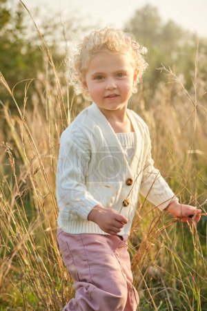 Téléchargez les photos : Adorable petite fille aux cheveux bouclés portant un pull blanc debout sur une pelouse herbeuse dans une campagne estivale ensoleillée et regardant la caméra - en image libre de droit