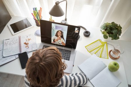 Photo for From above of kid having video call via netbook with girl playing flute at table with notebooks and modern tablet - Royalty Free Image