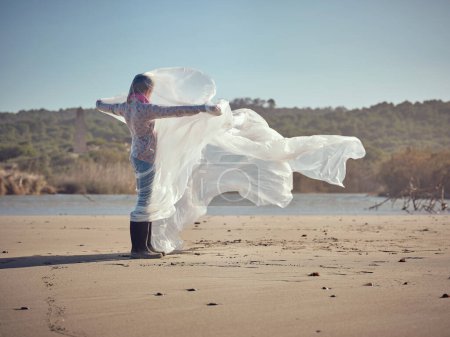 Téléchargez les photos : Vue latérale complète du corps de l'enfant debout sur une plage de sable près de la mer sereine et écartant les bras avec un film plastique ondulant - en image libre de droit