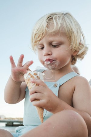 Foto de Desde abajo de adorable niño con el pelo rubio comiendo helado mientras se sienta en la playa durante las vacaciones de verano - Imagen libre de derechos