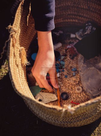 Photo for From above of crop anonymous person putting picked up piece of garbage into wicker basket with heap of various plastic wastes - Royalty Free Image
