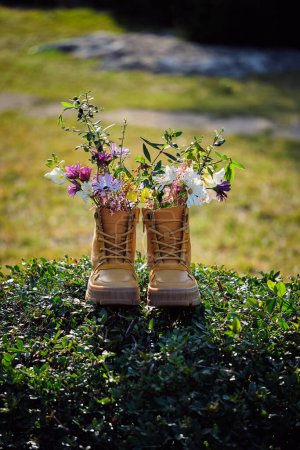 Foto de Par de botas marrones con flores silvestres de colores en el seto en el área rural con césped sobre fondo borroso durante el soleado día de verano - Imagen libre de derechos