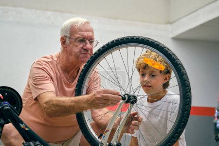 Téléchargez les photos : Attentif homme âgé dans les lunettes avec mignon petit-fils réparer roue de vélo moderne tout en passant du temps ensemble sur le parking - en image libre de droit