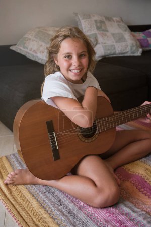 Téléchargez les photos : Enfant talentueux en vêtements décontractés assis sur le tapis près du coach et jouant de la musique à la guitare acoustique dans le salon à la maison le week-end - en image libre de droit