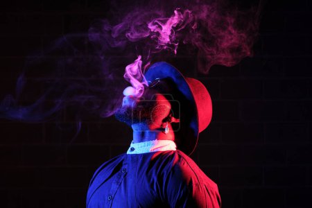 Photo for Side view of bearded man in stylish clothes and hat smoking cigarette while standing in dark colorful room - Royalty Free Image
