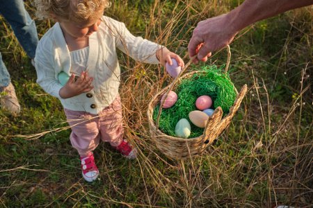 Foto de Desde arriba adorable niña en ropa casual poner coloridos huevos de Pascua en la cesta de mimbre en el césped cubierto de hierba en el campo soleado - Imagen libre de derechos
