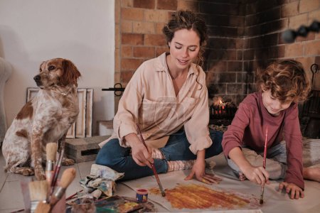 Photo for Young female artist in casual clothes and boy with brush painting on paper and looking down while spending time together at home near Spaniel dog - Royalty Free Image