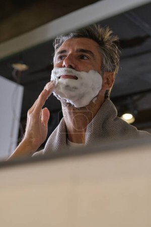 Photo for Low angle of bearded man looking at reflection in mirror and smiling while preparing for shaving in modern bathroom at home - Royalty Free Image