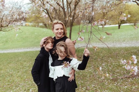 Téléchargez les photos : Femme heureuse et les enfants en tenues décontractées debout sous l'arbre en fleurs dans le parc et embrassant - en image libre de droit
