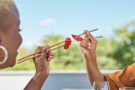 Photo for Side view of crop anonymous ethnic female friends holding tuna sashimi with wooden chopsticks together while having lunch in Asian restaurant - Royalty Free Image