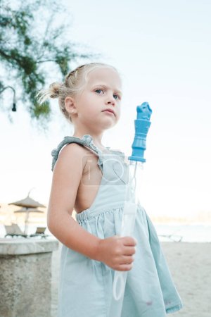 Photo for Side view of adorable little girl in dress holding plastic bottle of soap bubbles looking away at beach during summer vacation - Royalty Free Image