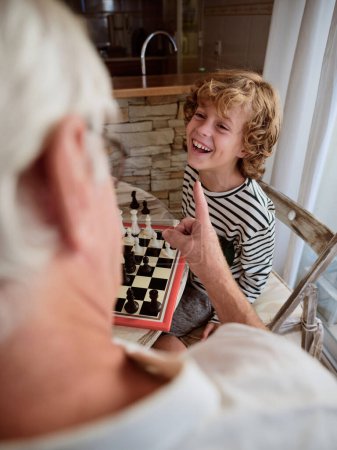 Foto de Chico positivo en ropa casual jugando al ajedrez con el abuelo anónimo de la cosecha mientras está sentado en la mesa en la cocina ligera en casa - Imagen libre de derechos