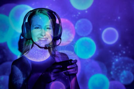 Photo for Happy young female with headset looking at camera with smile and pushing buttons on gamepad while playing videogame under colorful sports of light projection - Royalty Free Image