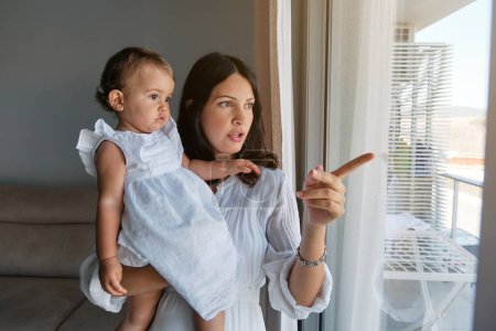 Foto de Mujer joven apuntando y sosteniendo al bebé curioso mientras exploran el mundo a través de la ventana juntos en la sala de estar en casa - Imagen libre de derechos