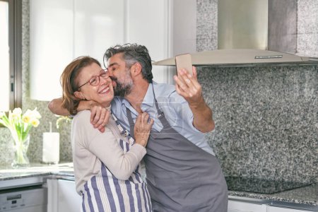 Téléchargez les photos : Heureux homme mûr et femme âgée positive dans des tabliers étreignant et s'amusant tout en prenant selfie dans la cuisine moderne - en image libre de droit
