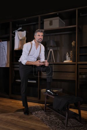 Photo for Full body of confident unshaven Hispanic man in elegant clothes looking away and using smartphone while standing against wooden shelves in atelier - Royalty Free Image