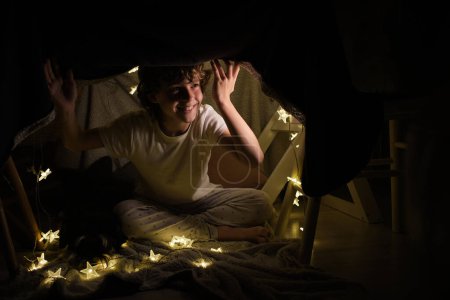 Photo for Full body of cheerful boy looking away while sitting under blanket construction with glowing garland in dark room at home - Royalty Free Image