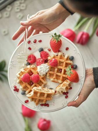 Photo for From above of crop anonymous person pouring honey on sweet baked waffles with ripe berries served on table in kitchen - Royalty Free Image