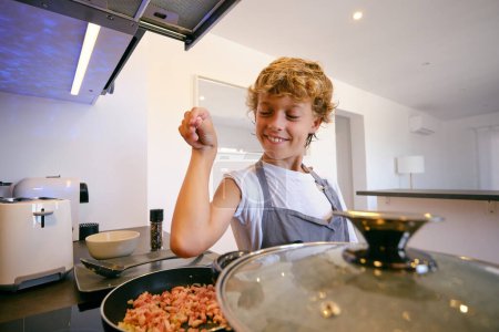 Foto de Niño sonriente en delantal rociando trozos de tocino ahumado con sal en la sartén en la estufa mientras cocina en casa - Imagen libre de derechos