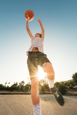 Téléchargez les photos : Angle bas du garçon en vêtements de sport lancer une balle orange tout en jouant au streetball contre le soleil sur le terrain de sport - en image libre de droit