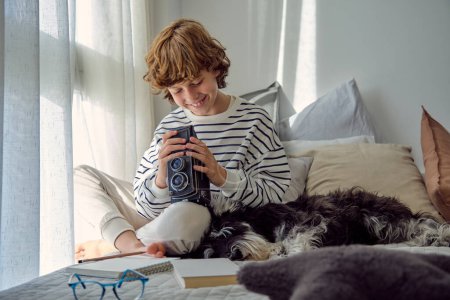 Photo for Cheerful schoolchild with vintage photo camera sitting with crossed legs near Miniature Schnauzer on bed in house - Royalty Free Image