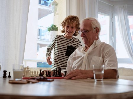 Téléchargez les photos : Petit-fils positif debout près de grand-père jouant jeu d'échecs intéressant à table tout en passant du temps ensemble dans le salon à la maison - en image libre de droit