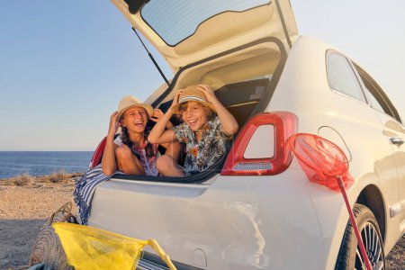 Photo for Two children sitting inside the trunk of the car making jokes while touching the straw hat. Baskets of objects for the beach next to the car with the sea in the background - Royalty Free Image