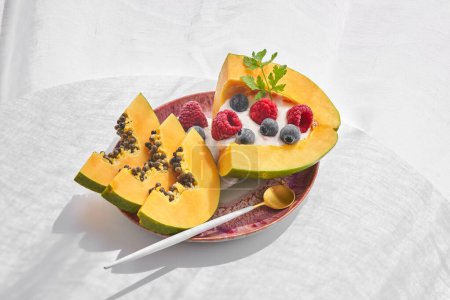 Foto de Porción de fruta dulce de papaya servida en plato de cerámica rosa con cuchara y decorada con yogur y bayas sobre fondo blanco - Imagen libre de derechos