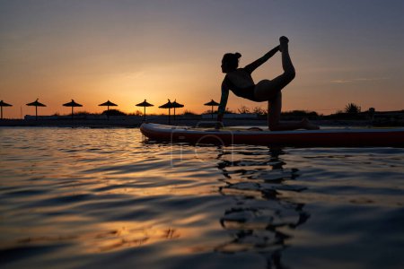 Téléchargez les photos : Corps complet d'athlète féminine un maillot de bain prenant Viaghrasana pose tout en pratiquant le yoga sur paddleboard au coucher du soleil - en image libre de droit