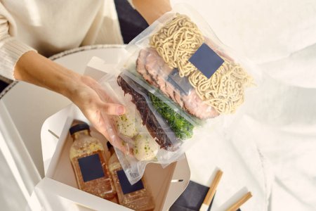 Téléchargez les photos : Personne anonyme de culture d'angle élevé montrant Ramen ingrédients de soupe emballés dans des sacs scellés sous vide au-dessus de la boîte à lunch - en image libre de droit