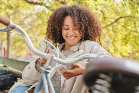 Foto de Mujer hispana sonriente con el pelo rizado en ropa casual sentada en el banco cerca de la bicicleta y descansando mientras lee interesante libro durante el fin de semana en el parque - Imagen libre de derechos
