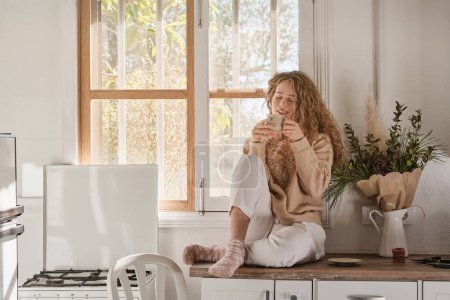 Téléchargez les photos : Corps complet de jeune femme en vêtements décontractés assis sur la garniture de cuisine et boire une boisson chaude dans la chambre à la maison - en image libre de droit