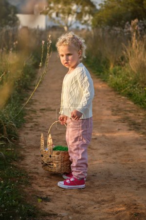 Foto de Vista lateral adorable niña en ropa elegante que lleva canasta de mimbre y de pie en estrecha carretera rural en la naturaleza verde - Imagen libre de derechos