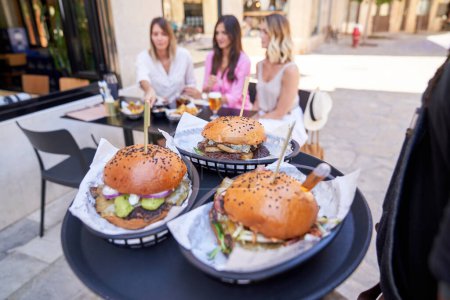 Téléchargez les photos : Serveur méconnaissable portant plateau avec des hamburgers savoureux pour les femmes assises à la table ensemble et parlant en attendant l'ordre dans le café de trottoir - en image libre de droit