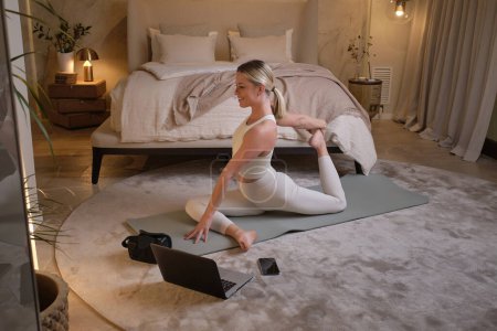 Foto de Cuerpo completo de atleta femenina en ropa deportiva estirando las piernas y sentada en Pigeon asana mientras practica yoga sobre estera en el dormitorio con portátil en casa - Imagen libre de derechos