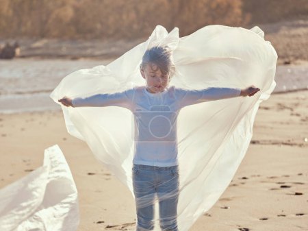 Téléchargez les photos : Préadolescent paisible debout avec les mains étendues enveloppé dans un film plastique ramassé sur le rivage sablonneux contaminé de prospérer dans la journée ensoleillée - en image libre de droit