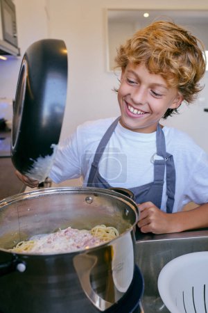 Foto de Alegre niño con sartén añadiendo salsa cremosa a la pasta sabrosa en olla mientras se cocina en casa - Imagen libre de derechos