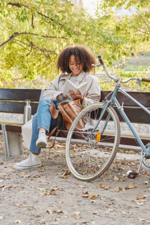 Foto de Cuerpo completo de joven estudiante sonriente de etnia femenina con el pelo rizado sentado en el banco en el parque cerca de la bicicleta y libro de lectura mientras hace la tarea - Imagen libre de derechos